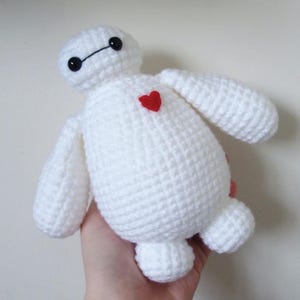 May include: A handmade white crochet plush toy with black eyes and a red heart on its chest. The toy has a rounded body, arms, and legs, resembling a cartoon character. The plush is held in a hand against a plain background.