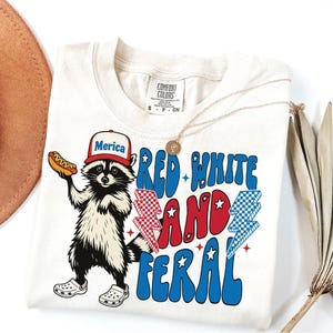 May include: Cream-colored t-shirt featuring a raccoon wearing a baseball cap with the word "Merica" and holding a hot dog. The shirt has the text "Red White and Feral" in red and blue, with star and lightning bolt graphics. A gold necklace is draped on the shirt.