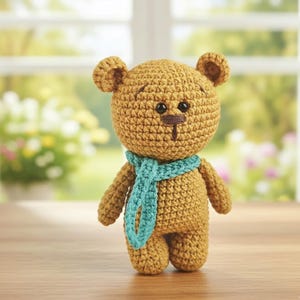 May include: A handmade crochet teddy bear in shades of tan and brown, wearing a teal scarf. The bear has small black eyes and a brown stitched nose and mouth. The toy is standing in front of a window with a blurred outdoor background.