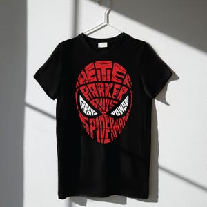 Puede incluir: Camiseta negra con un diseño rojo de la máscara de Spider-Man. El diseño de la máscara está formado por las palabras "Peter Parker", "Great Power" y "Spider-Man". La camiseta está colgada de una percha blanca.
