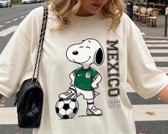 Puede incluir: Una camiseta de manga corta color hueso con el gráfico de un perro de dibujos animados que viste una camiseta de fútbol verde y pantalones cortos blancos, con un pie sobre un balón. El texto "MEXICO" y "2026" aparece impreso verticalmente.