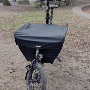 Könnte beinhalten: Ein schwarzes Lastenrad mit einem großen Frontkorb, der von einer schwarzen Plane bedeckt ist. Das Fahrrad hat einen schwarzen Rahmen, einen Lenker und ein einzelnes Vorderrad. Das Fahrrad steht auf einem Feldweg.