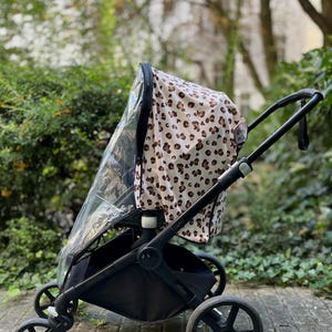 Könnte beinhalten: Ein schwarzer Kinderwagen mit einem Leopardenmuster-Verdeck und einer transparenten Regenhülle. Der Kinderwagen hat vier Räder und einen Aufbewahrungskorb. Der Hintergrund zeigt grünes Laub und einen gepflasterten Weg.