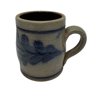 Può includere: Una tazza in gres con interno scuro e manico. La tazza presenta un esterno marrone chiaro maculato con un motivo a foglie blu scuro. Una fascia blu scuro circonda la parte superiore della tazza.