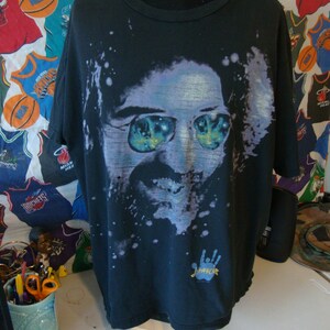 Puede incluir: Camiseta negra con una imagen descolorida de un hombre con gafas de sol. La imagen est&aacute; rodeada de pintura blanca salpicada. La camiseta tiene una huella de mano azul con la palabra "Jimi" escrita en amarillo.