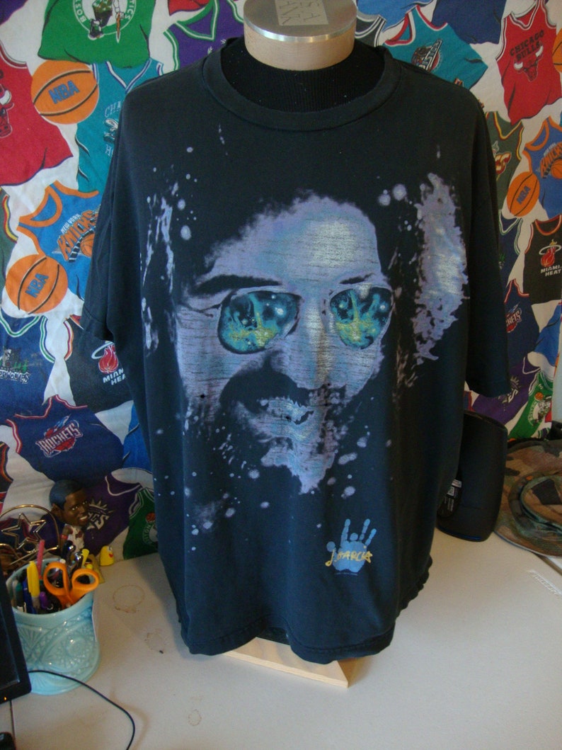 Puede incluir: Camiseta negra con una imagen descolorida de un hombre con gafas de sol. La imagen est&aacute; rodeada de pintura blanca salpicada. La camiseta tiene una huella de mano azul con la palabra "Jimi" escrita en amarillo.