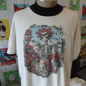 May include: White t-shirt with a black ringer neck featuring a colorful graphic of a skeleton with roses and a skull. The graphic is surrounded by a decorative frame.