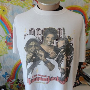 以下が含まれることがあります： 3人のミュージシャンのグラフィックが特徴の白いTシャツ。「63rd Annual Hampton Jazz Festival」の文字が赤色でプリントされています。デザインには音符とレトロなフォントが含まれています。