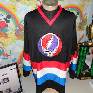 May include: Black long-sleeve jersey with a red V-neck and striped sleeves. The jersey features a large circular emblem with a skull and lightning bolt design in red, purple, and white. The jersey is displayed on a mannequin.