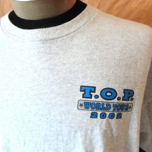 May include: Light gray T-shirt with a black collar. The shirt features the blue text "T.O.P. WORLD TOUR 2002" on the front. The shirt is made of a soft material and has short sleeves.