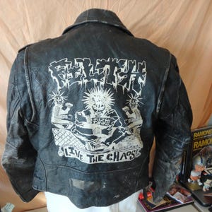 Op de afbeelding: Zwarte leren jas met een witte afbeelding op de rug. De afbeelding toont het woord "FILTH" in een gestileerd lettertype, illustraties en de zin "LIVE THE CHAOS". De jas heeft een klassieke biker-stijl.