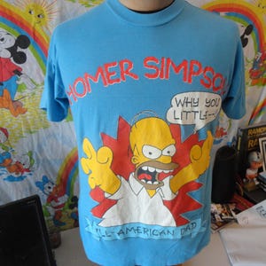 May include: A light blue t-shirt featuring Homer Simpson with the text "HOMER SIMPSON" in red. Homer is depicted with an angry expression, arms raised, and the phrase "WHY YOU LITTLE--!" in a speech bubble. The shirt also says "ALL-AMERICAN DAD".