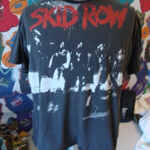 May include: Black t-shirt with a white and gray image of the band Skid Row. The band's name is printed in red letters across the top of the shirt.