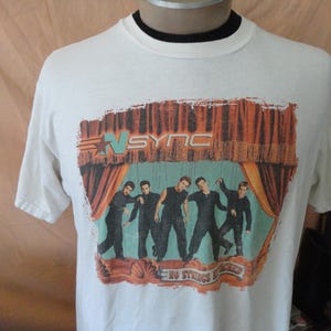 May include: White t-shirt featuring a graphic of the band *NSYNC on a stage with a red curtain backdrop. The band members are depicted in black outfits. The text "*NSYNC" and "NO STRINGS ATTACHED" are also visible.