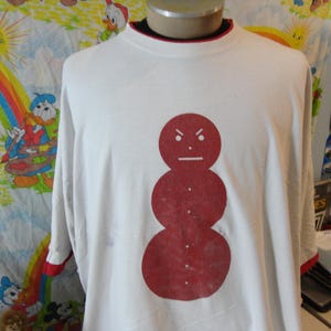 May include: White long-sleeve t-shirt with a red snowman graphic. The snowman has an angry face and is composed of three circles with small white dots. The shirt has a red trim on the sleeves and collar.