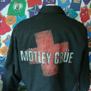 Op de afbeelding: Zwarte jas met een rood kruisontwerp en de bandnaam "Mötley Crüe" in witte letters. De jas is donker van kleur en lijkt gemaakt van een duurzaam materiaal. Het ontwerp staat op de achterkant van de jas.