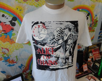 Puede incluir: Camiseta blanca con un diseño gráfico en blanco y negro y las palabras "ALICE IN CHAINS" en rojo. El diseño incluye un reloj con una cara de luna y figuras de un cuento. La camiseta se muestra en un maniquí.