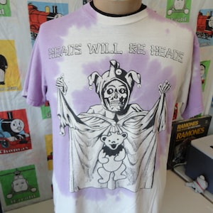 May include: A purple tie-dye t-shirt with a white graphic of a jester holding a teddy bear. The text "Heads Will Be Heads" is printed in black on the shirt.