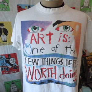 May include: White t-shirt with a colorful graphic print of an eye and the text "ART is: One of the FEW THINGS LEFT WORTH doing".
