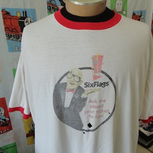 Könnte beinhalten: Weißes T-Shirt mit einem rot-weißen Ringeldesign. Das Shirt zeigt eine Grafik eines Mannes im Smoking, der ein Schild mit der Aufschrift "Six Flags" hält. Der Text "Ask me about your ticket to fun!" ist unter der Grafik gedruckt.