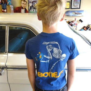 May include: Blue t-shirt with a skateboarding skeleton graphic and the word "BONES" in yellow. The shirt is short-sleeved and worn by a person. A white car is in the background.