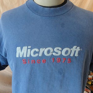 Może przedstawiać: Niebieski t-shirt z krótkim rękawem z białym logo Microsoft i czerwonym napisem "Since 1975". Koszulka ma okrągły dekolt i wydaje się być wykonana z miękkiego, wygodnego materiału.