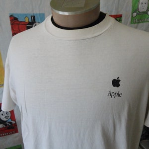 以下が含まれることがあります： 黒いAppleのロゴとテキストが入ったオフホワイトのTシャツ。Tシャツはクルーネックで半袖です。背景には、電車のキャラクターが描かれたカラフルな生地があります。