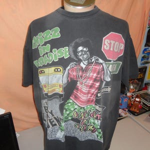 May include: Dark gray t-shirt with a graphic of a person wearing glasses and a red and white striped shirt. The shirt has the words "Whizz in Paradise" and "Stop" on it. The person is wearing gold chains and green and white patterned pants.