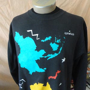 May include: Black crew neck sweatshirt with a colorful map design. The map features blue, yellow, red, and white accents, with the text "Apple Pacific" in white. A white zig-zag and star are also present.