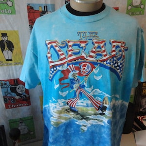 Könnte beinhalten: Ein blaues Batik-T-Shirt mit einem Motiv von Onkel Sam, der eine Friedensflagge hält. Das Motiv ist von Sternen und Streifen umgeben. Der Text "THE DEAD" ist in großen Buchstaben auf dem Shirt gedruckt.