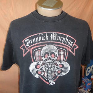 May include: A black short-sleeve t-shirt featuring a graphic print of three skulls arranged around a central spine. Above the skulls, a red and white banner displays the text Dropkick Murphys in a gothic font, set against a plaid shield.
