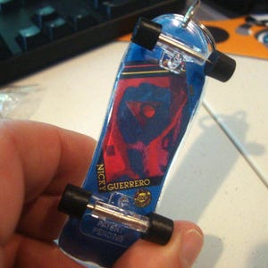 Puede incluir: Un llavero de monopatín en miniatura con un diseño azul y rojo. El llavero tiene ruedas negras y ejes plateados. El nombre "NICKY GUERRERO" está impreso en el lateral, junto con "PATENT PENDING".