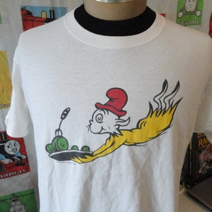 May include: White t-shirt with a cartoon illustration of a cat wearing a red hat and flying through the air with a plate of green peas. The text "The Cat in the Hat" is printed below the image.