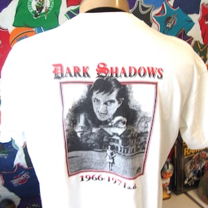 Puede incluir: Una camiseta blanca de manga corta con un estampado gráfico en la espalda. El estampado muestra el texto Dark Shadows en rojo y negro, una ilustración en blanco y negro de un vampiro y una mansión, y el texto 1966-1971 a.d. en la parte inferior.