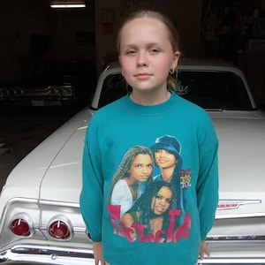 May include: Teal sweatshirt featuring a graphic of three women with the text "The Tlc Tlc Tlc". The sweatshirt is worn by a person standing in front of a white car.