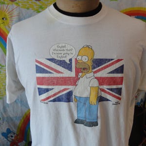 May include: White t-shirt featuring a cartoon image of Homer Simpson in front of a distressed Union Jack flag. Homer is wearing a white shirt and blue pants. Text in a speech bubble reads, "English! What reads that? I'm never going to England!"