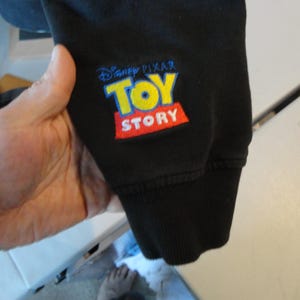 Może przedstawiać: Czarna bluza z haftowanym logo "Disney Pixar Toy Story" w kolorze ż&oacute;łtym, niebieskim i czerwonym.