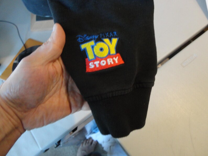 Może przedstawiać: Czarna bluza z haftowanym logo "Disney Pixar Toy Story" w kolorze ż&oacute;łtym, niebieskim i czerwonym.