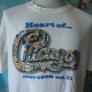 May include: White t-shirt with a blue and white graphic of the word "Chicago" made up of smaller images. The text "Heart of..." is above the graphic and the text "1967-1995 Vol. II" is below the graphic.