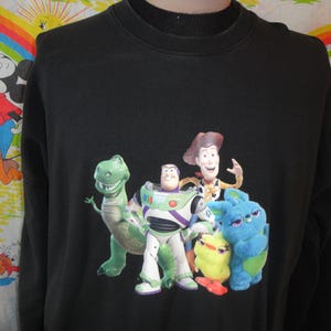 Może przedstawiać: Czarna bluza z długim rękawem z nadrukiem postaci z serii Toy Story. Postacie to Woody, Buzz Astral, Rex i fioletowy kosmita.