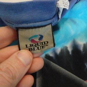 May include: A close-up of a Liquid Blue brand tag on a blue and turquoise tie-dye shirt. The tag is brown with a white border and the text "Liquid Blue" in white.