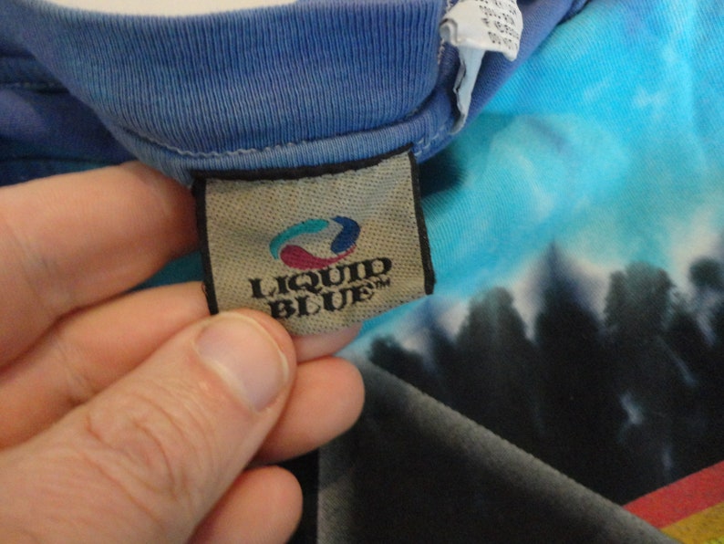 May include: A close-up of a Liquid Blue brand tag on a blue and turquoise tie-dye shirt. The tag is brown with a white border and the text "Liquid Blue" in white.
