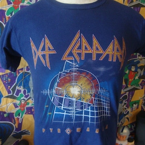 Könnte beinhalten: Ein blaues T-Shirt mit einem Def Leppard-Logo und dem Schriftzug "Pyromania" auf der Vorderseite.