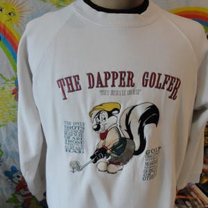 Könnte beinhalten: Weißer Langarm-Pullover mit einem Cartoon-Stinktier, das eine Golfmütze trägt und einen Golfschläger hält. Der Text "The Dapper Golfer" ist in Rot auf der Vorderseite des Pullovers aufgedruckt. Der Text "Tous Jours Le Course" ist in kleineren roten Buchstaben unter dem Haupttext aufgedruckt.