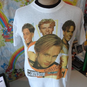 May include: White t-shirt featuring a graphic of four young men. The shirt has the words "CAUGHT" and German text. The shirt is displayed on a mannequin.