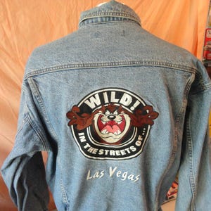 May include: A light blue denim jacket featuring a large circular embroidered patch on the back. The patch depicts the Tasmanian Devil character with the text WILD! IN THE STREETS OF... and the embroidered white text Las Vegas below it.