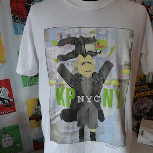 Puede incluir: Camiseta blanca con un diseño de Kermit la Rana y Miss Piggy en Nueva York. El diseño incluye el texto "KP NYC" en verde. La camiseta es de cuello redondo.