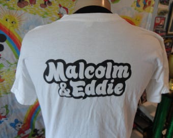 Pode incluir: Camiseta branca com o texto "Malcolm & Eddie" em uma fonte de bolha preta e branca. A camisa é exibida em um manequim. O fundo inclui um lençol estampado colorido e outros itens.