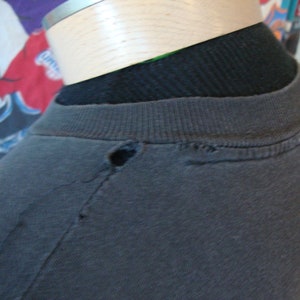 May include: A black t-shirt with a small hole near the neckline. The shirt is on a mannequin.