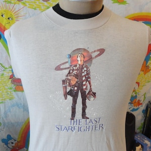 May include: White t-shirt with a graphic print of a man holding a weapon, standing in front of a planet with rings. The text "The Last Starfighter" is printed below the graphic.